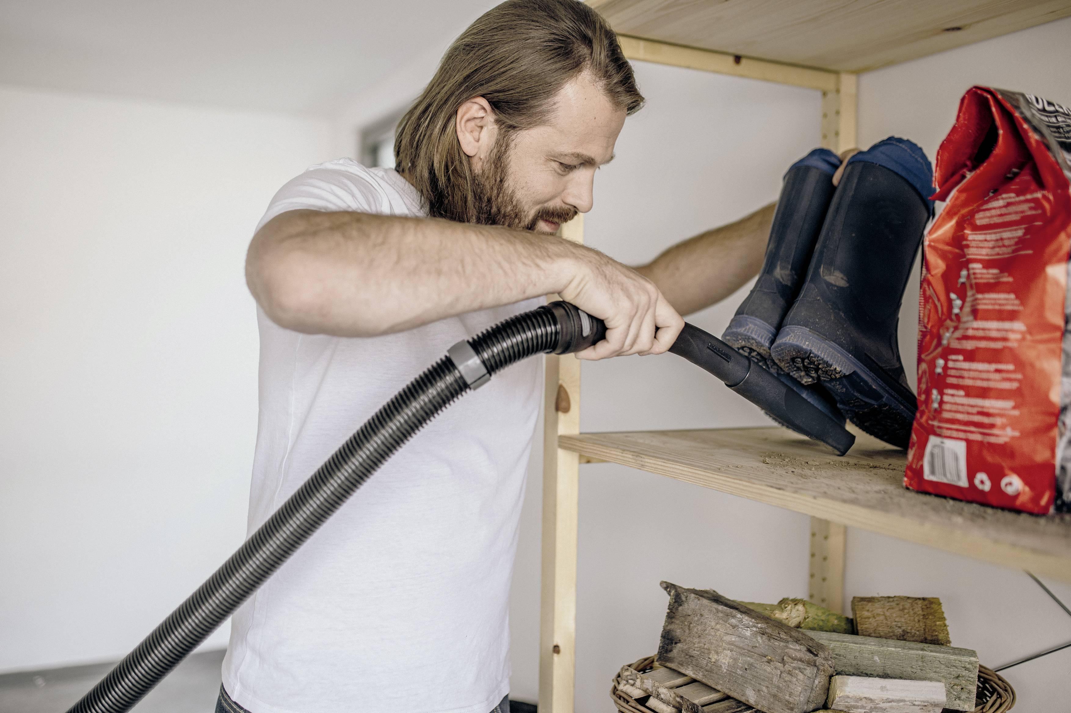 A man with a beard is cleaning a shelf in the storage room with a vacuum cleaner. Next to him are blue wellington boots and a feed sack.