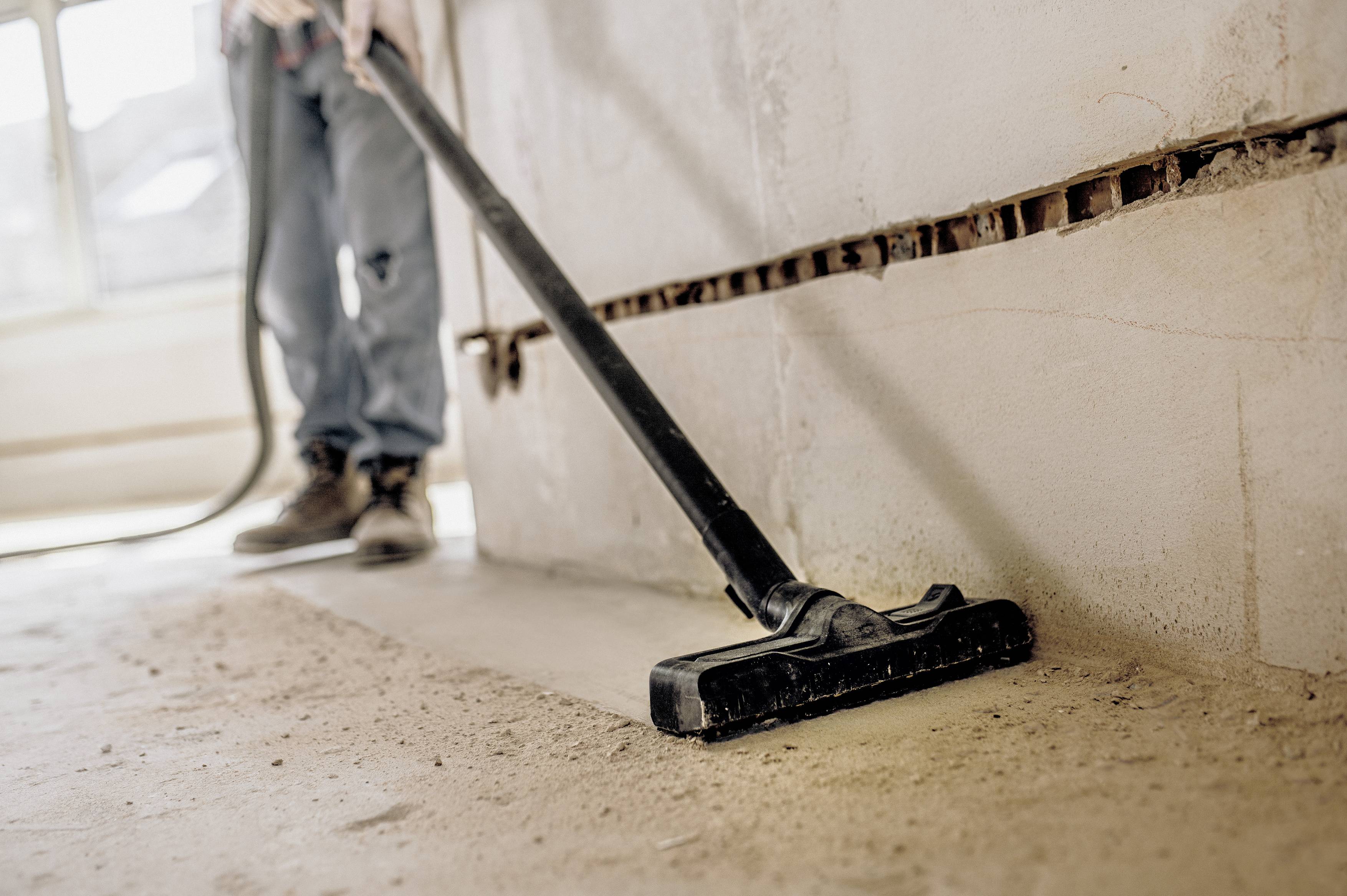 A person is cleaning a dusty floor with a vacuum cleaner in an unfinished room. Bare brick walls are visible in the background.