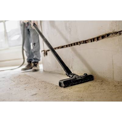 A person is cleaning a dusty floor with a vacuum cleaner in an unfinished room. Bare brick walls are visible in the background.