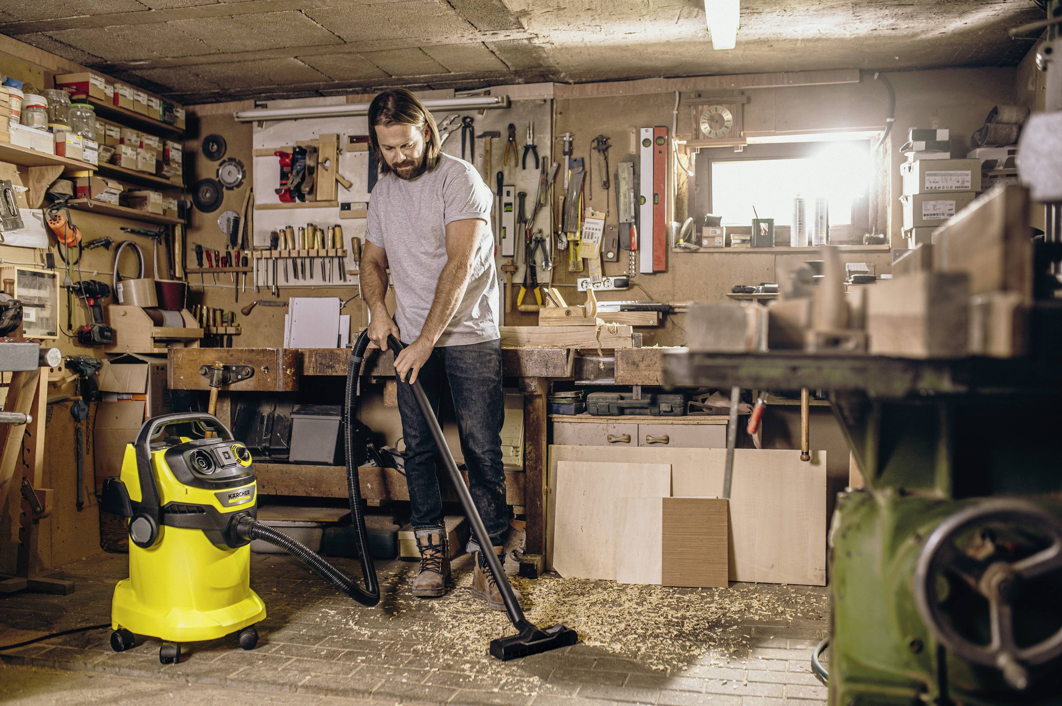 A man is vacuuming sawdust with a workshop vacuum cleaner on the floor of a well-equipped workshop.
