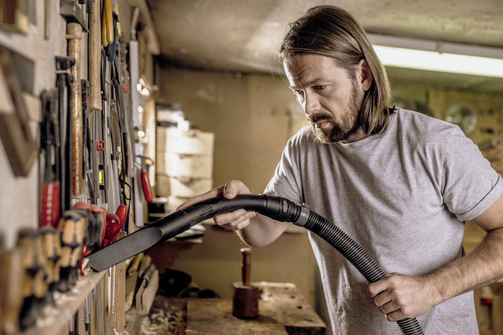 A person using a vacuum to clean tools hanging on a workshop wall. The space is organized with various hand tools visible.