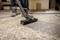 A person wearing work boots uses a vacuum cleaner to clean sawdust off a concrete floor, suggesting a workshop or construction setting.