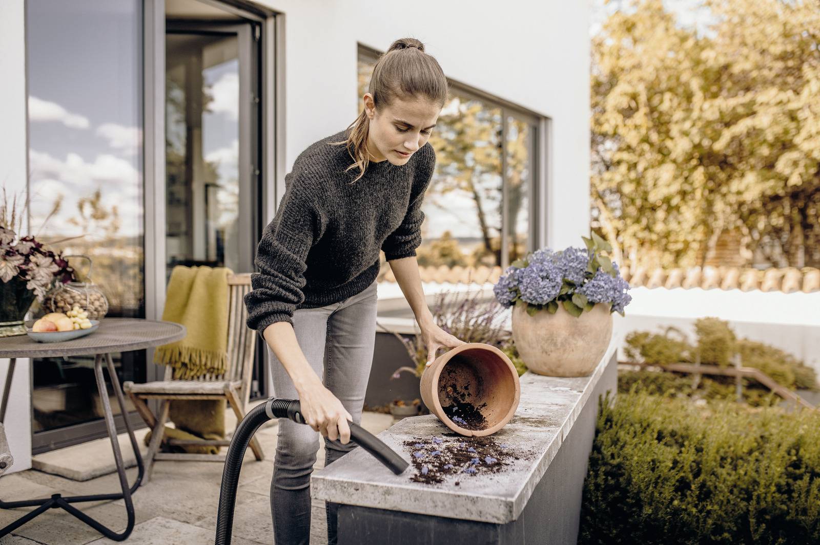 A person is vacuuming soil from a table on a patio, with potted flowers and a chair nearby, under a clear sky.