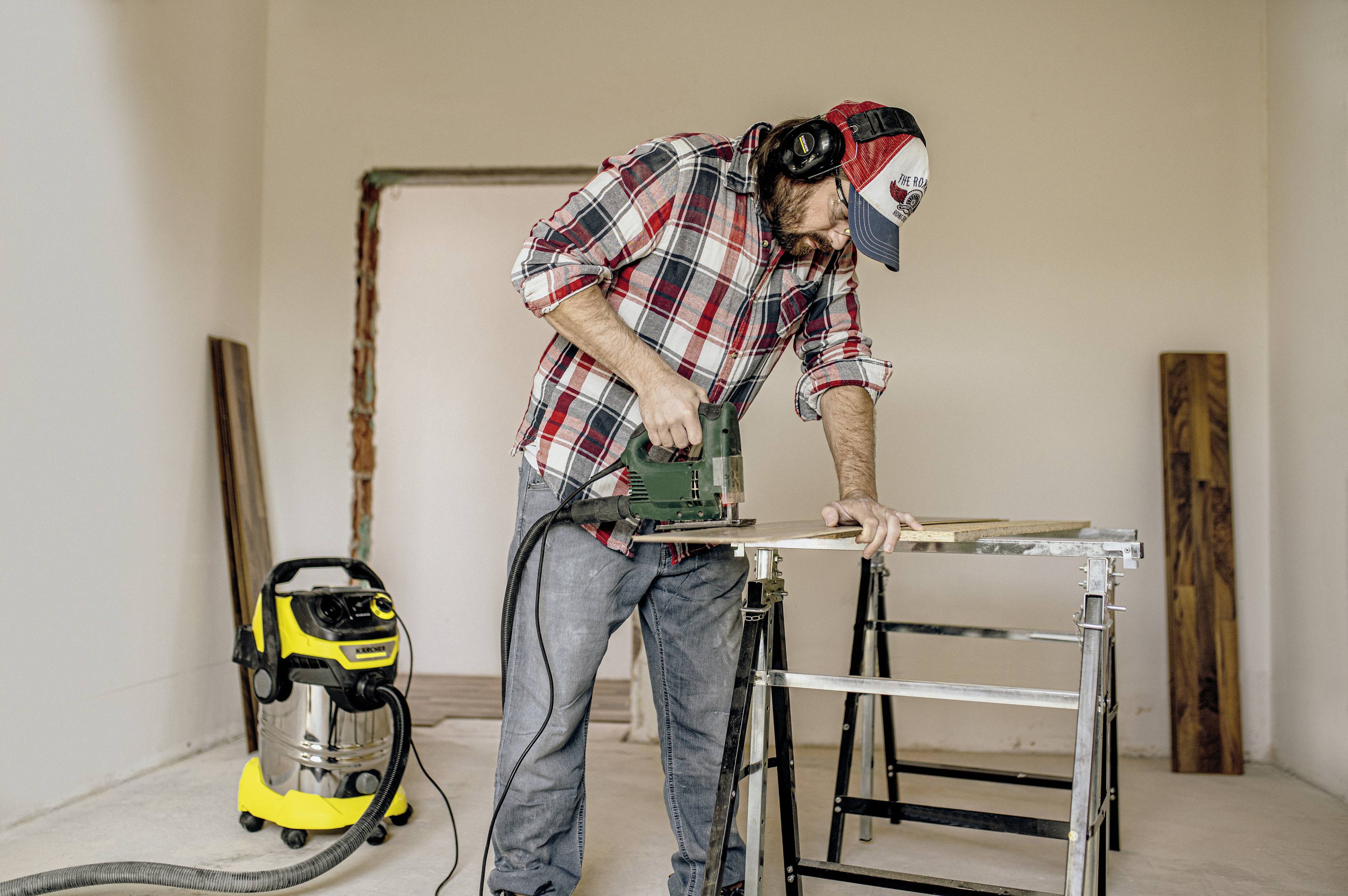 A man is wearing a lumberjack shirt and a cap, working with an electric jigsaw on a piece of wood. A vacuum cleaner is standing in the background.