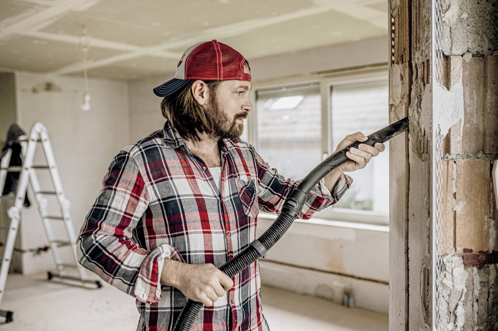 Man in a plaid shirt and red cap cleaning a wooden beam with a vacuum in a room under renovation.