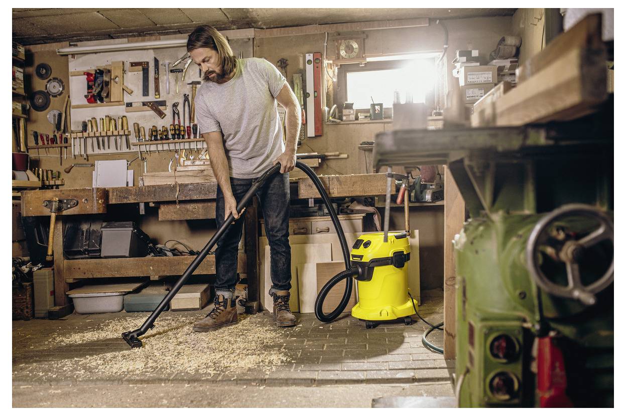 A person in a workshop using a vacuum to clean wood shavings off the floor. Various tools and a fully stocked workbench are visible.