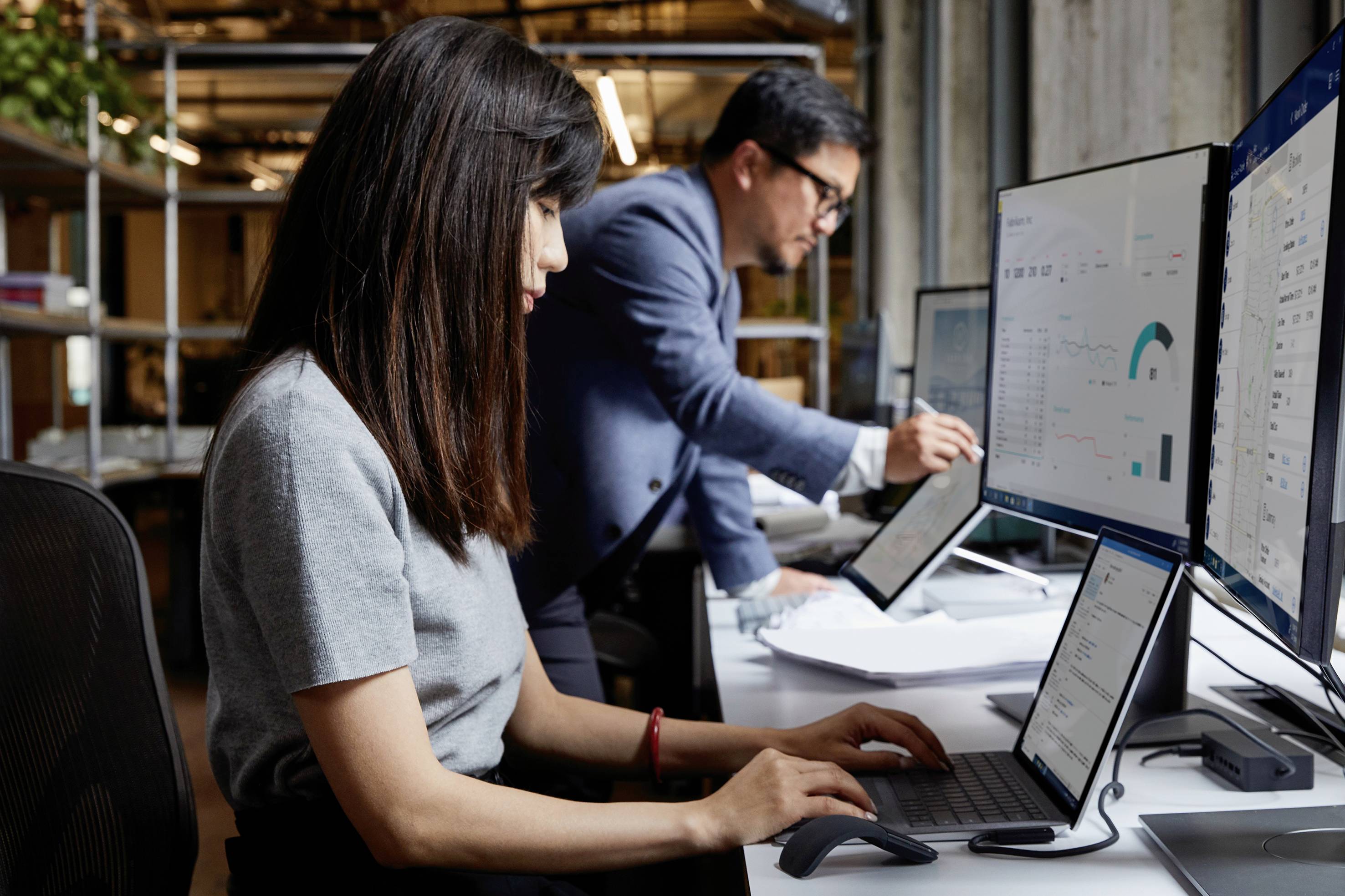 Two people are working at computer workstations. In the foreground, a woman is using a laptop, with a man in the background working on a tablet, surrounded by monitors.