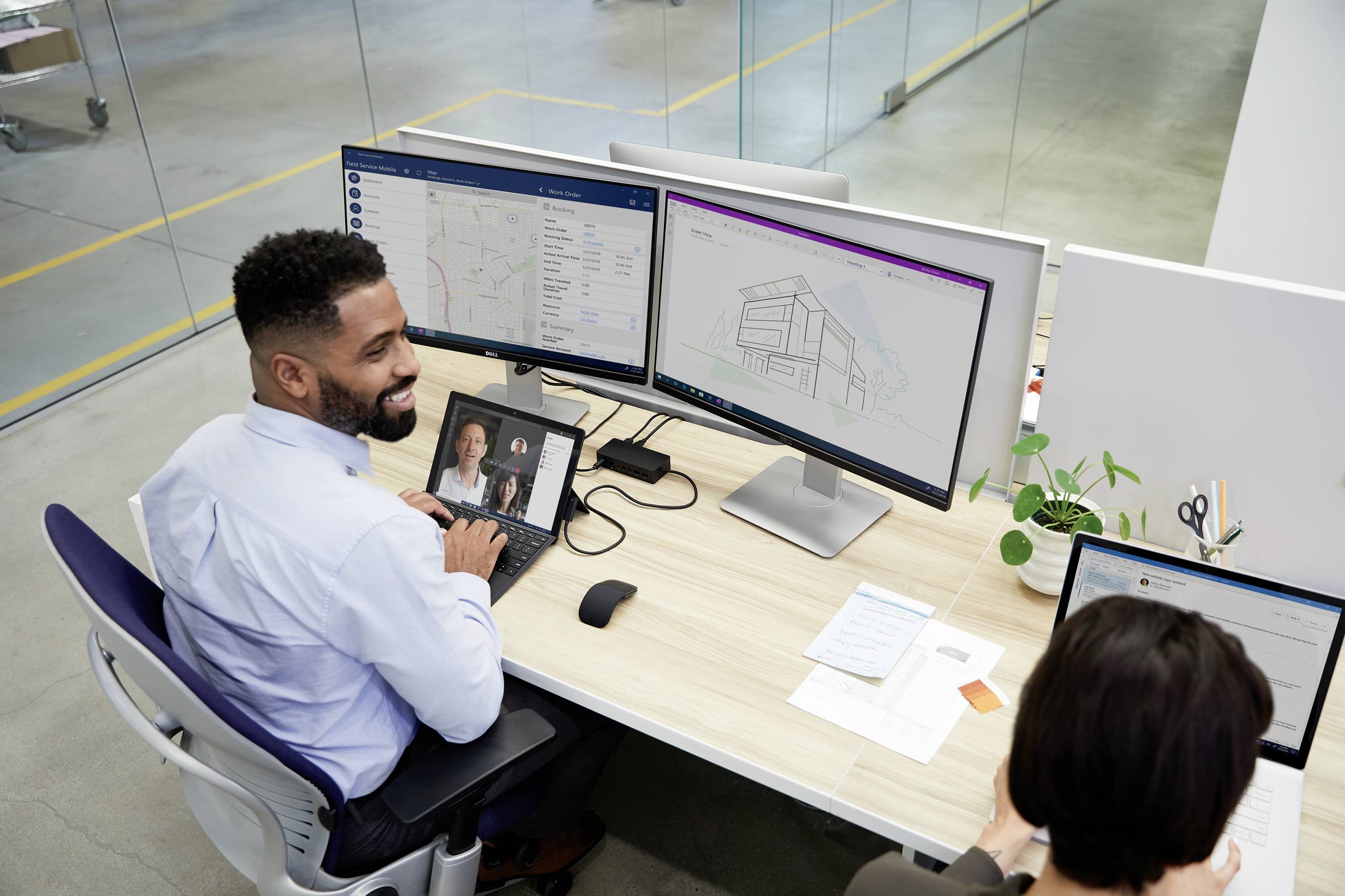 A man sits at a desk with multiple monitors and smiles. A video call with two people is visible on one of the screens.