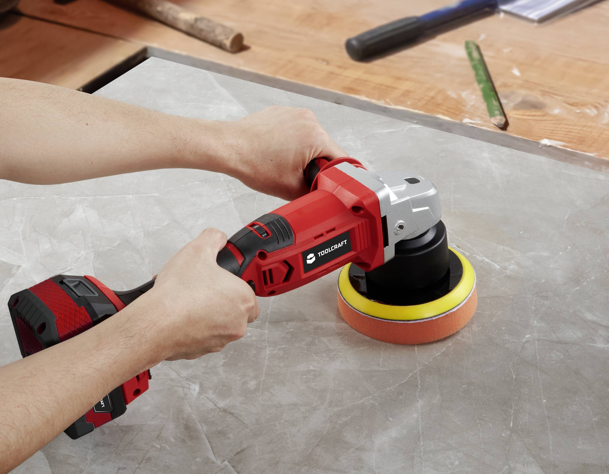 A person is using a red, electric polishing device on a grey stone surface. A table with tools is in the background.
