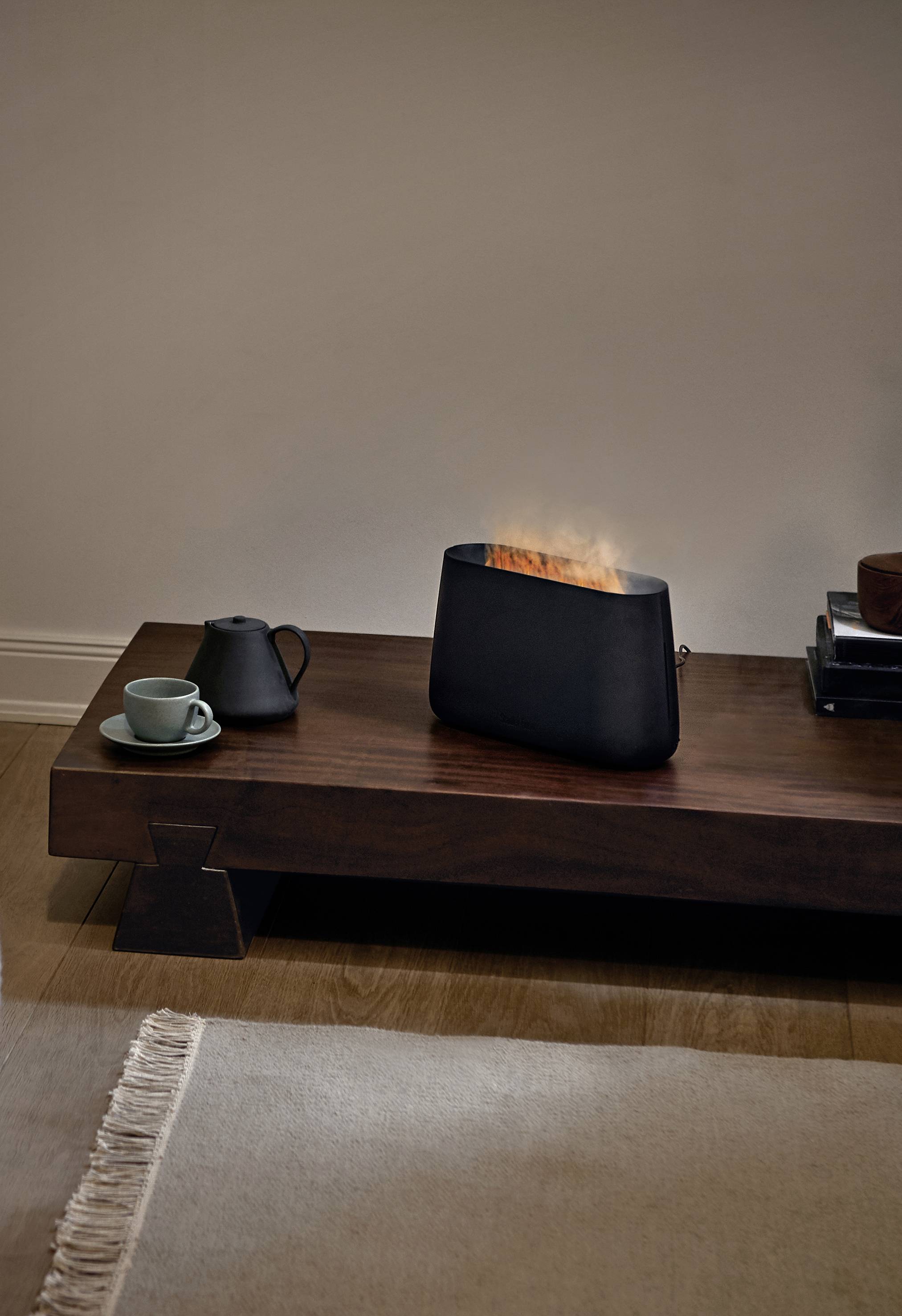 A contemporary fireplace on a wooden table, beside it a black teapot and a cup. The fireplace creates a soft, simulated flame.