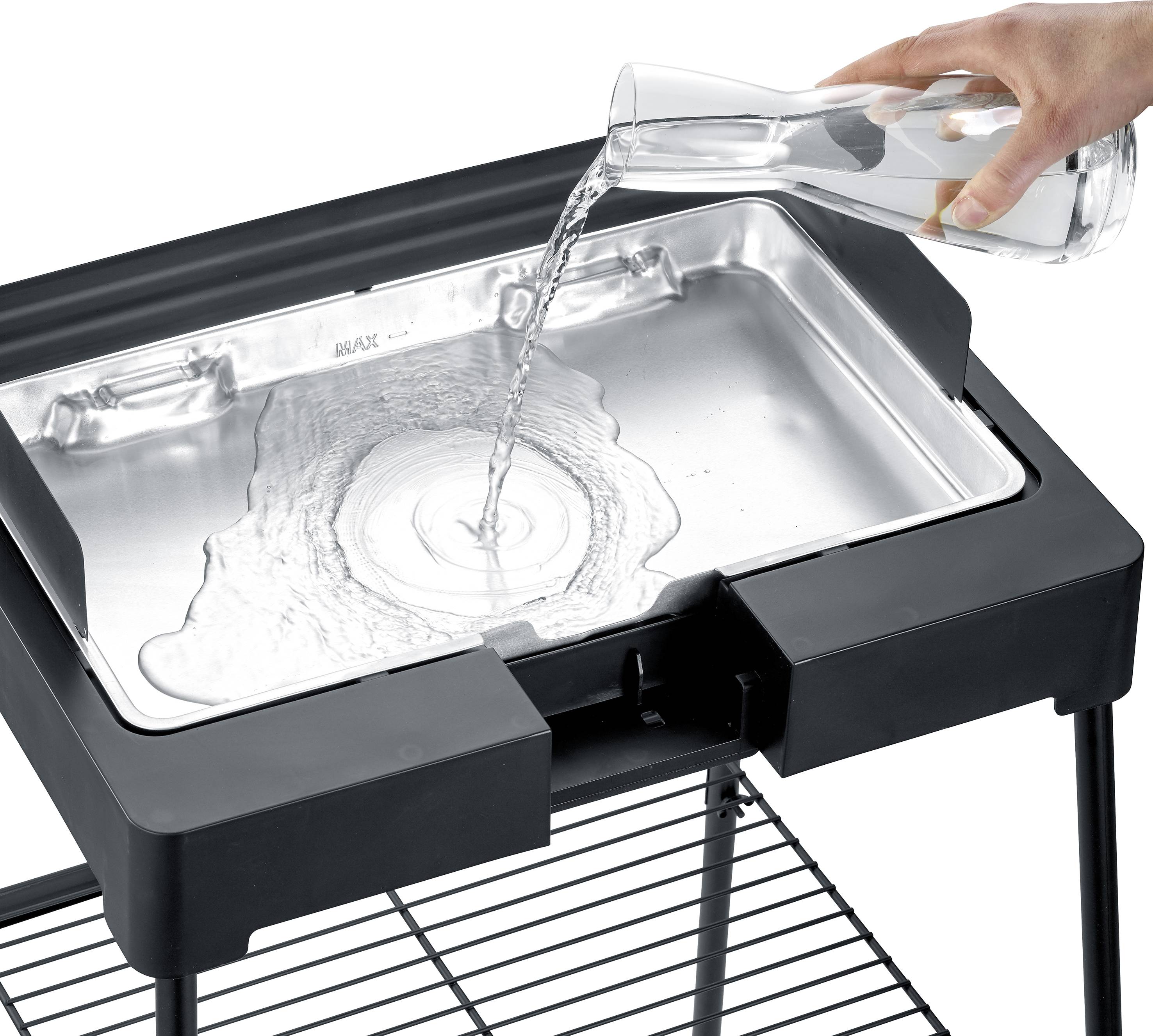 A hand is pouring water from a glass carafe into a rectangular metal tray on a black barbecue trolley.