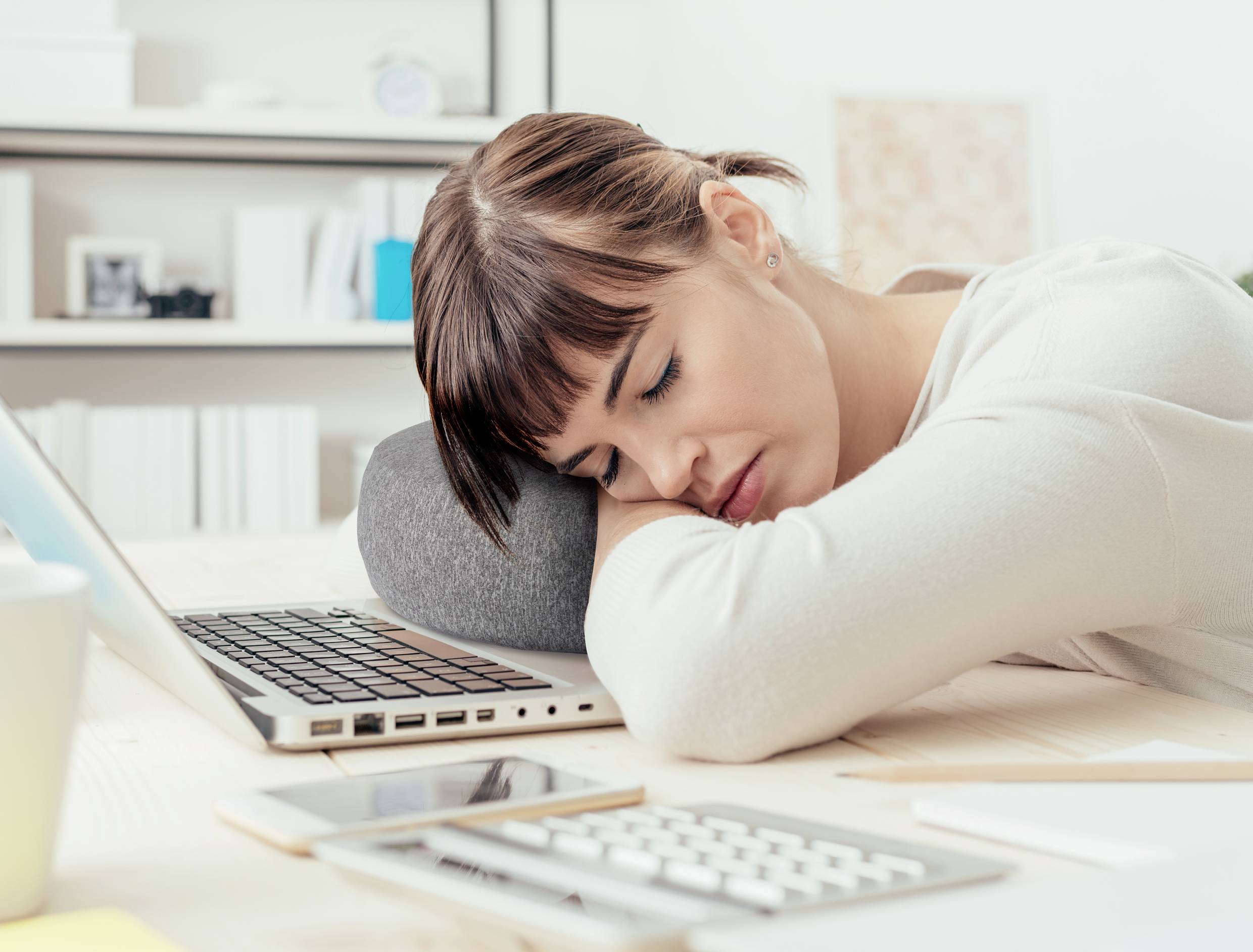 A woman is sleeping with her head on a desk next to a laptop, symbolising fatigue at work.