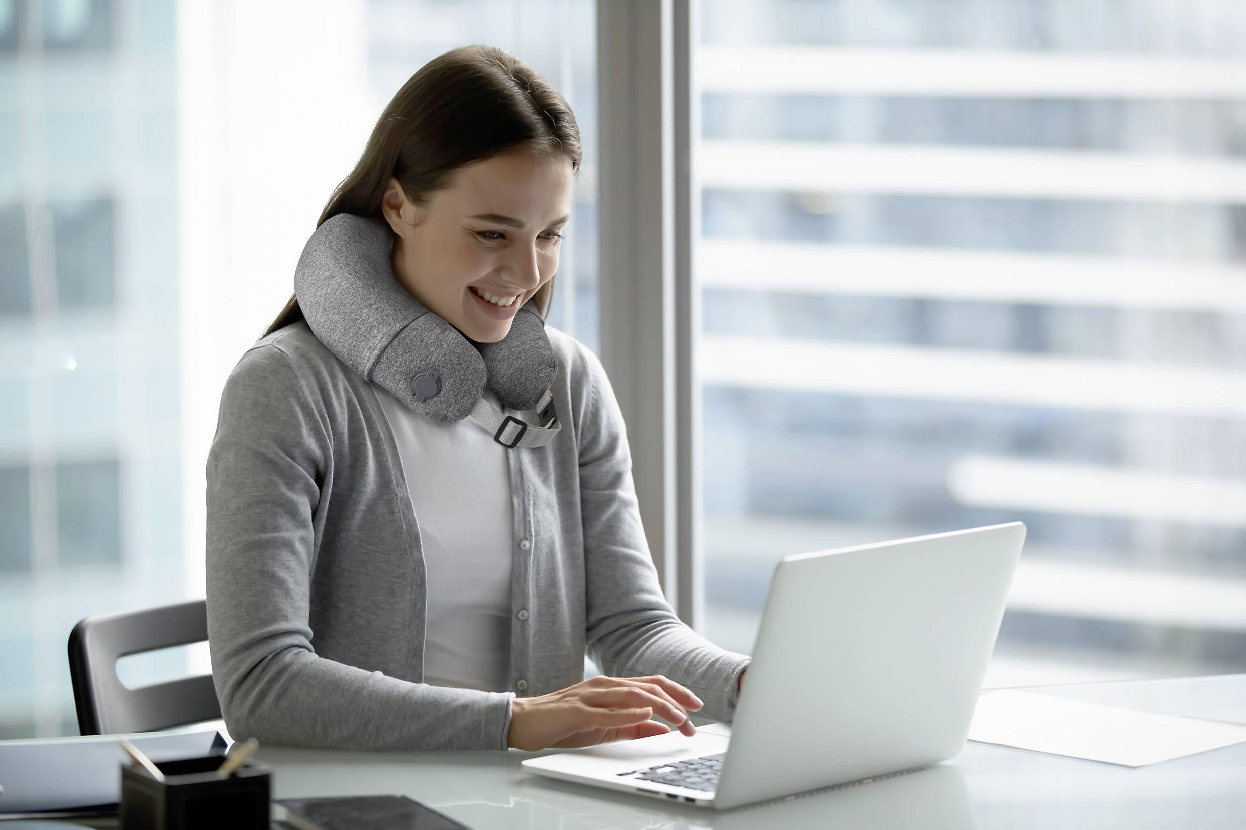 A smiling woman at a laptop is wearing a neck pillow in a modern office.