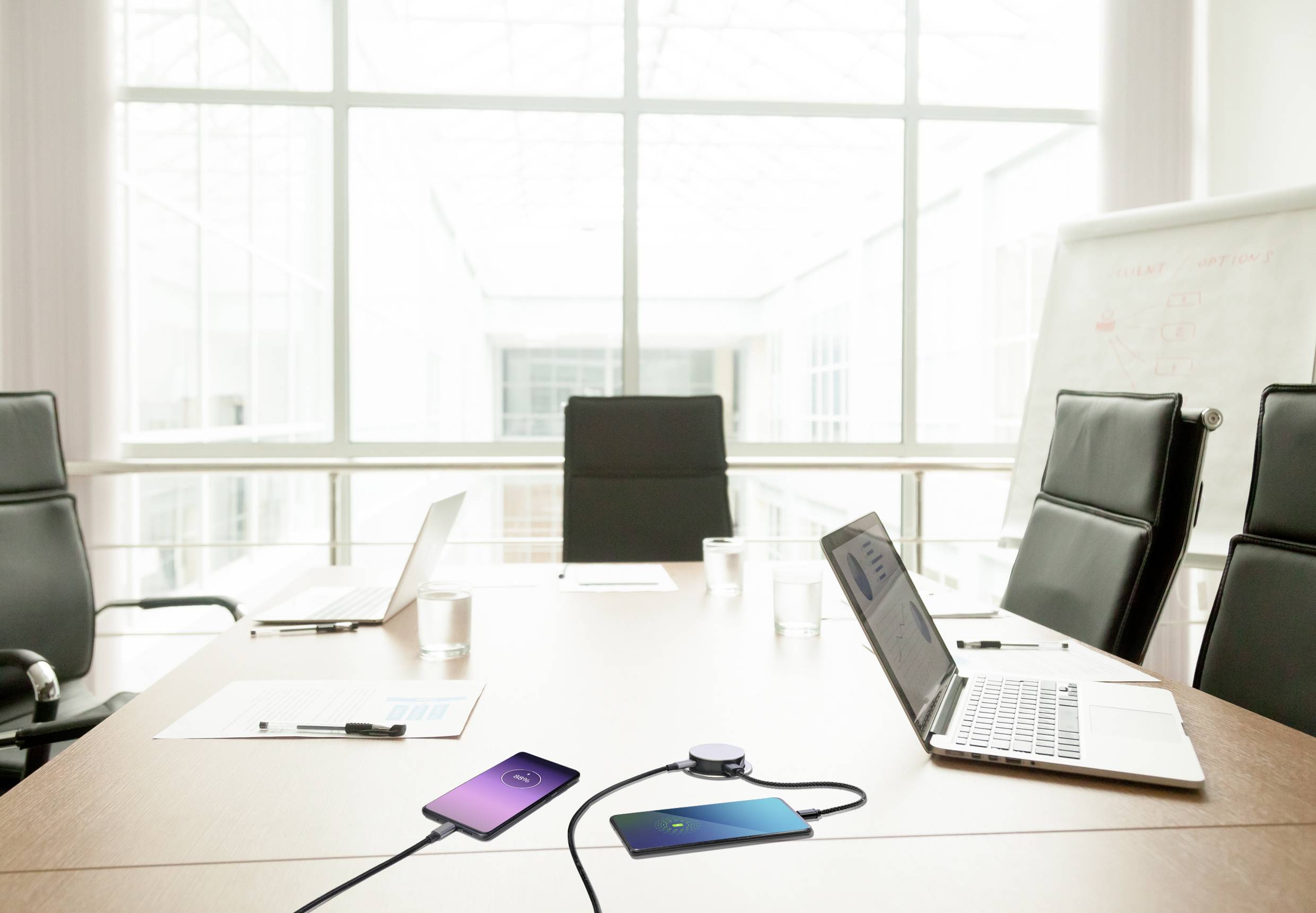A conference room with a table, on which two laptops and two smartphones are connected to a charger. A whiteboard is in the background.