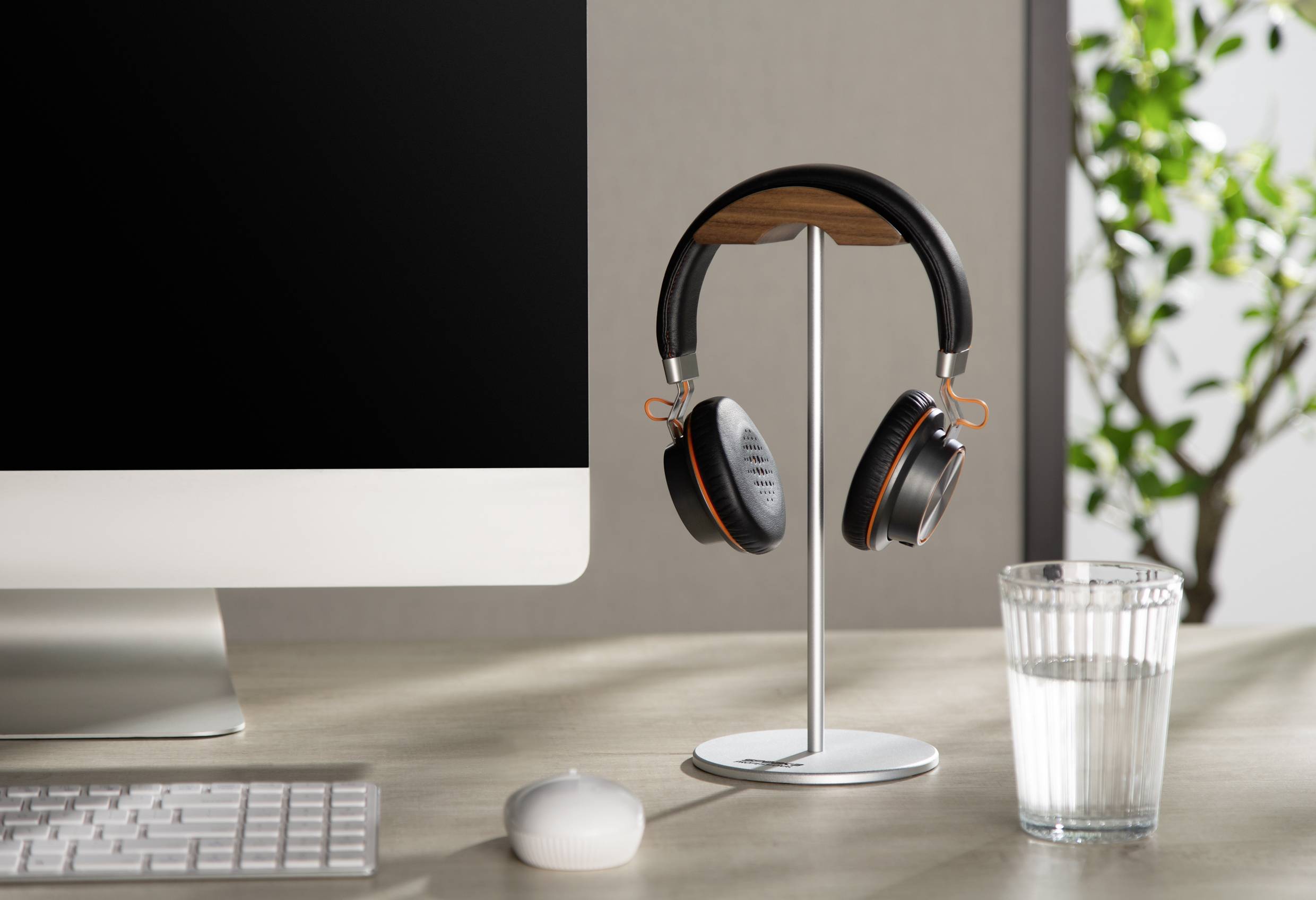 'Desk with monitor, wireless headphones on stand, glass of water, white mouse and keyboard. Plant in the background.'