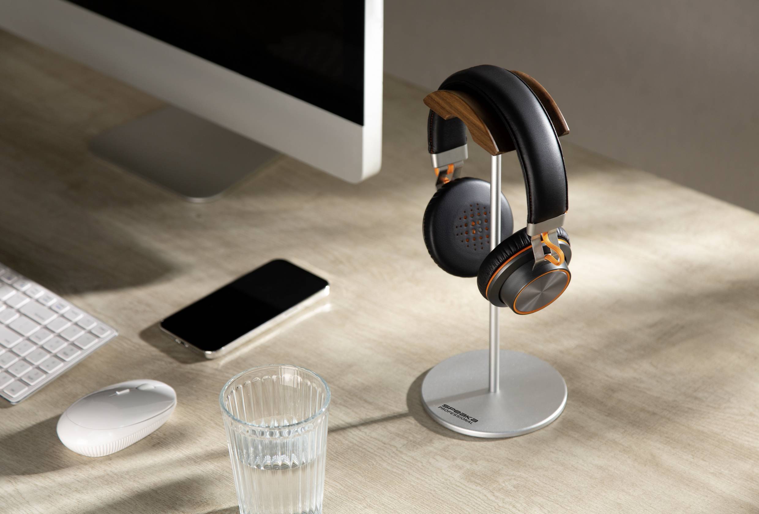 On a desk sit a computer monitor, a wireless keyboard and mouse, a glass and headphones on a stand.