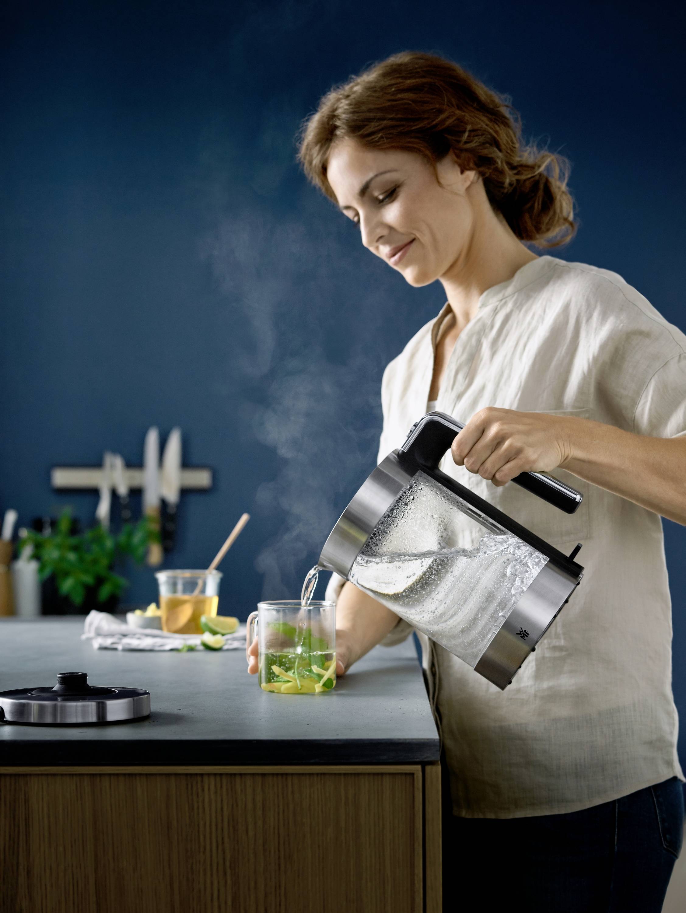 A woman pours boiling water from an electric kettle into a cup with herbs, steam rising. Kitchen in the background with blue walls.