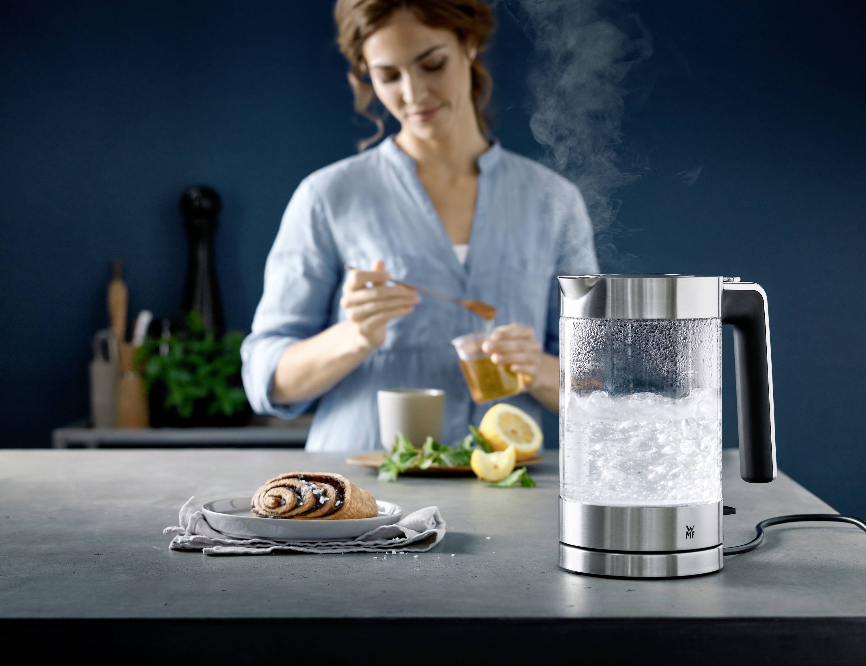 A woman is preparing tea with hot water from a kettle; a pastry lies in the foreground.