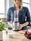 Woman in blue blouse using a blender to make a fruit smoothie. Fresh berries and yoghurt on the table. Kitchen in the background.