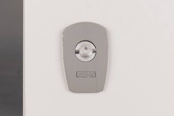 A grey wall light with a circular lens in the centre, mounted on a white wall. It serves to provide illumination.