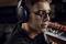 A man wearing glasses and headphones is looking intently at something. Keyboards are blurrily visible in the background.