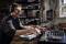 A person wearing headphones sits at a table filled with music production equipment, working intently, with a laptop open.
