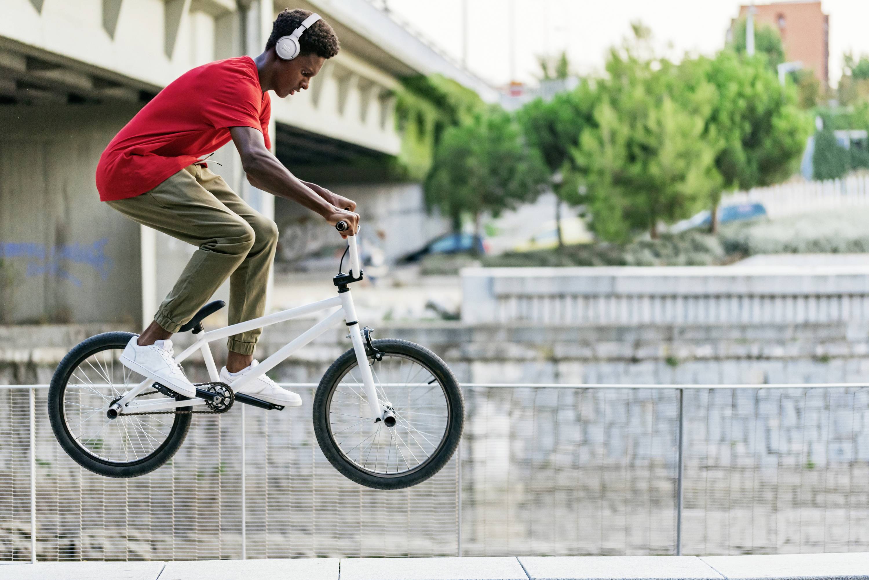 A young man in a red T-shirt and headphones is riding a BMX bike along a railing. Trees and a bridge are visible in the background.