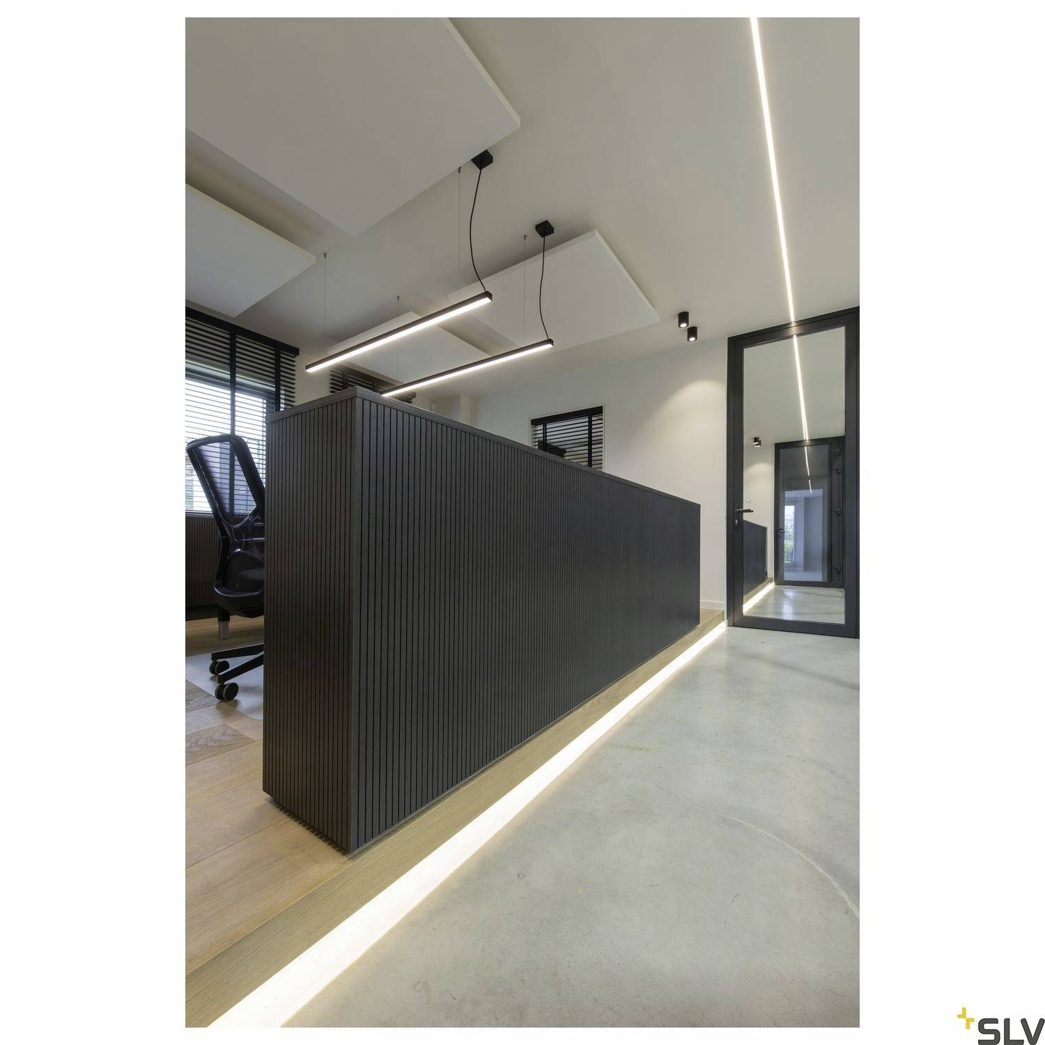 A modern office with a large, dark reception desk. The room has light-coloured walls, a glass partition, lamps, and a wooden floor.