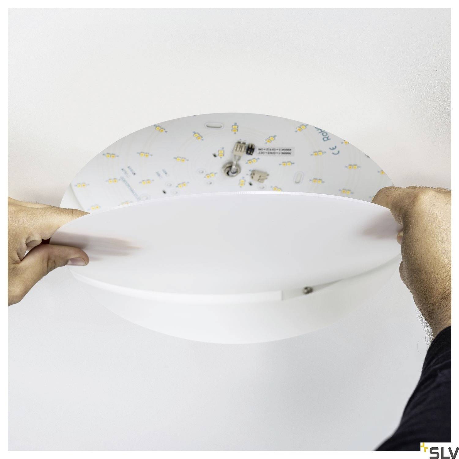 A person is installing a round LED ceiling light. Hands are holding the light fixture while attaching it to the ceiling.