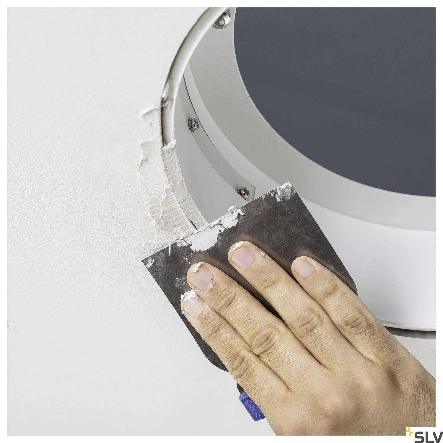A hand smooths white plaster around a round ceiling opening with a plastering trowel.