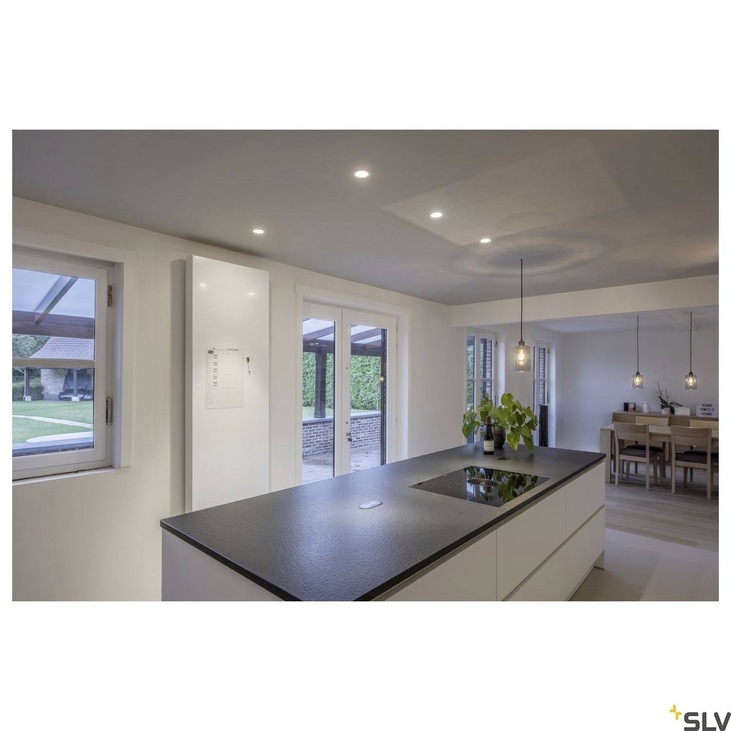 Modern kitchen with an island and dining area, bright lighting, large windows with an external view, minimalist interior design.