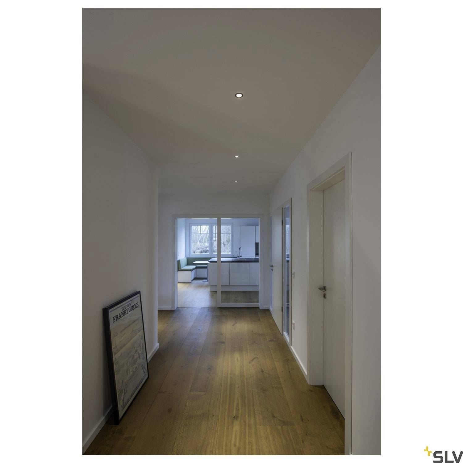 Corridor with a wooden floor, white walls, and ceiling lights. A framed picture is leaning against the wall on the left. At the end, there is a door leading to the kitchen.