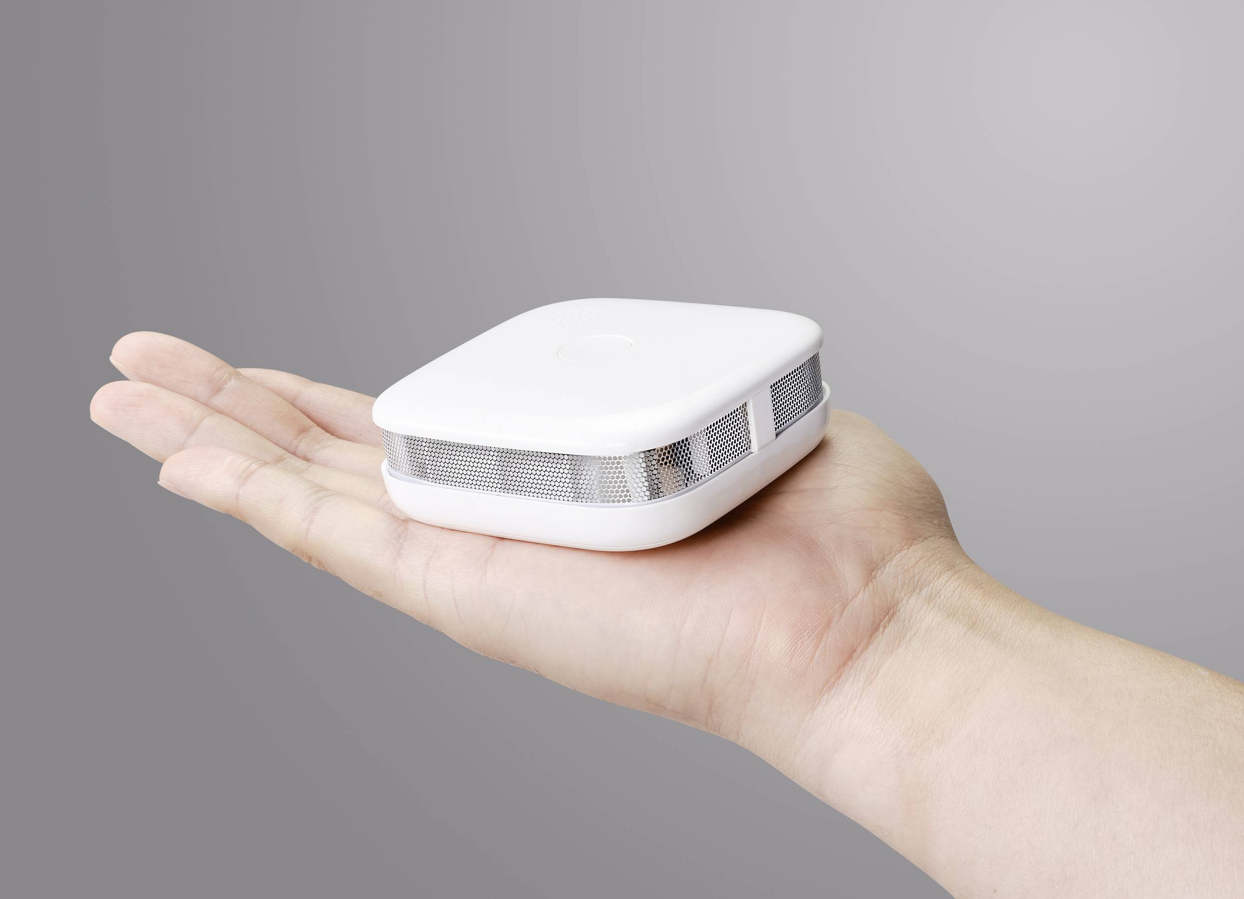 A hand is holding a small, square, white device with ventilation slots, against a grey background.