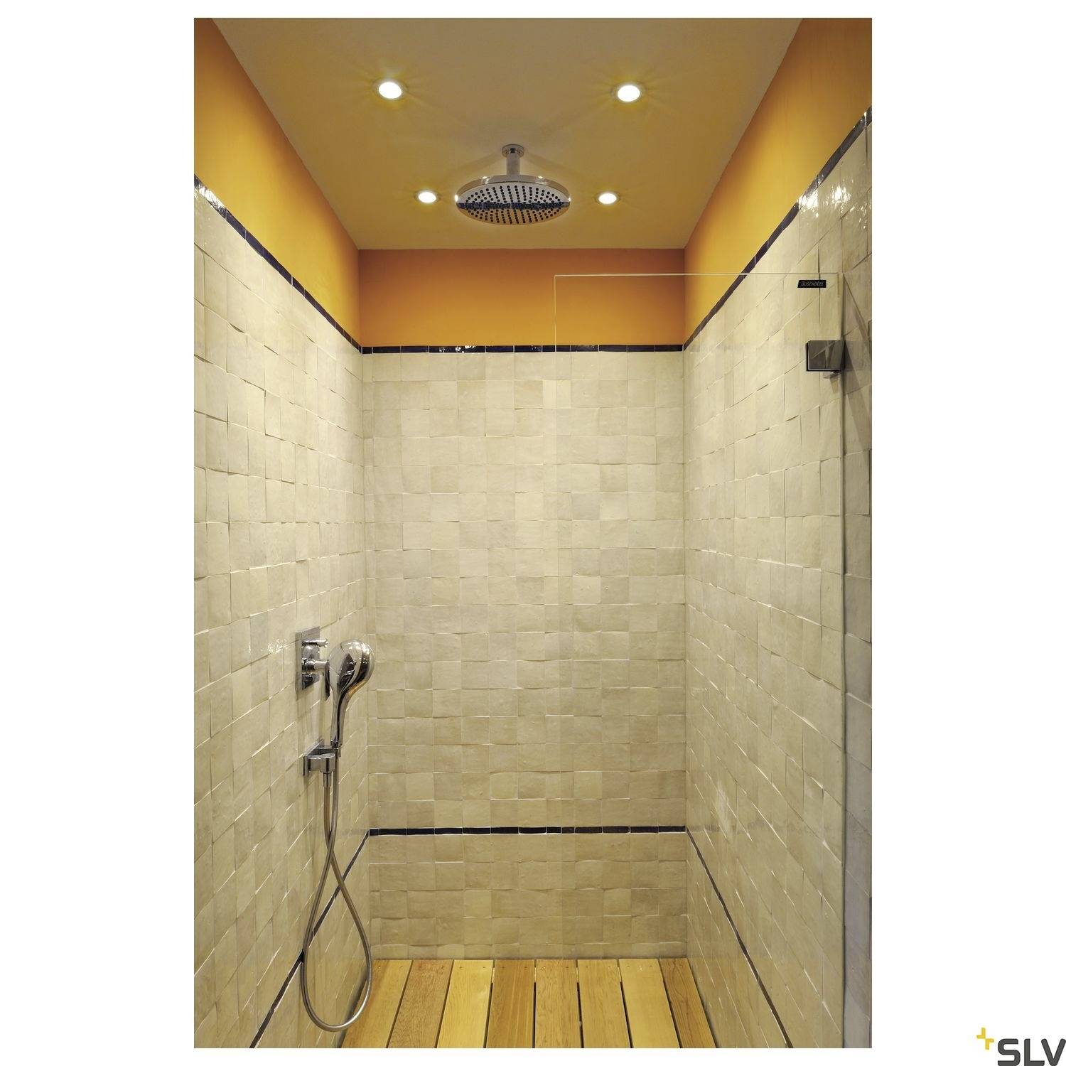 This image shows a shower with beige tiled walls and a wooden shower mat. A ceiling-mounted shower head and a wall-mounted shower head are visible.