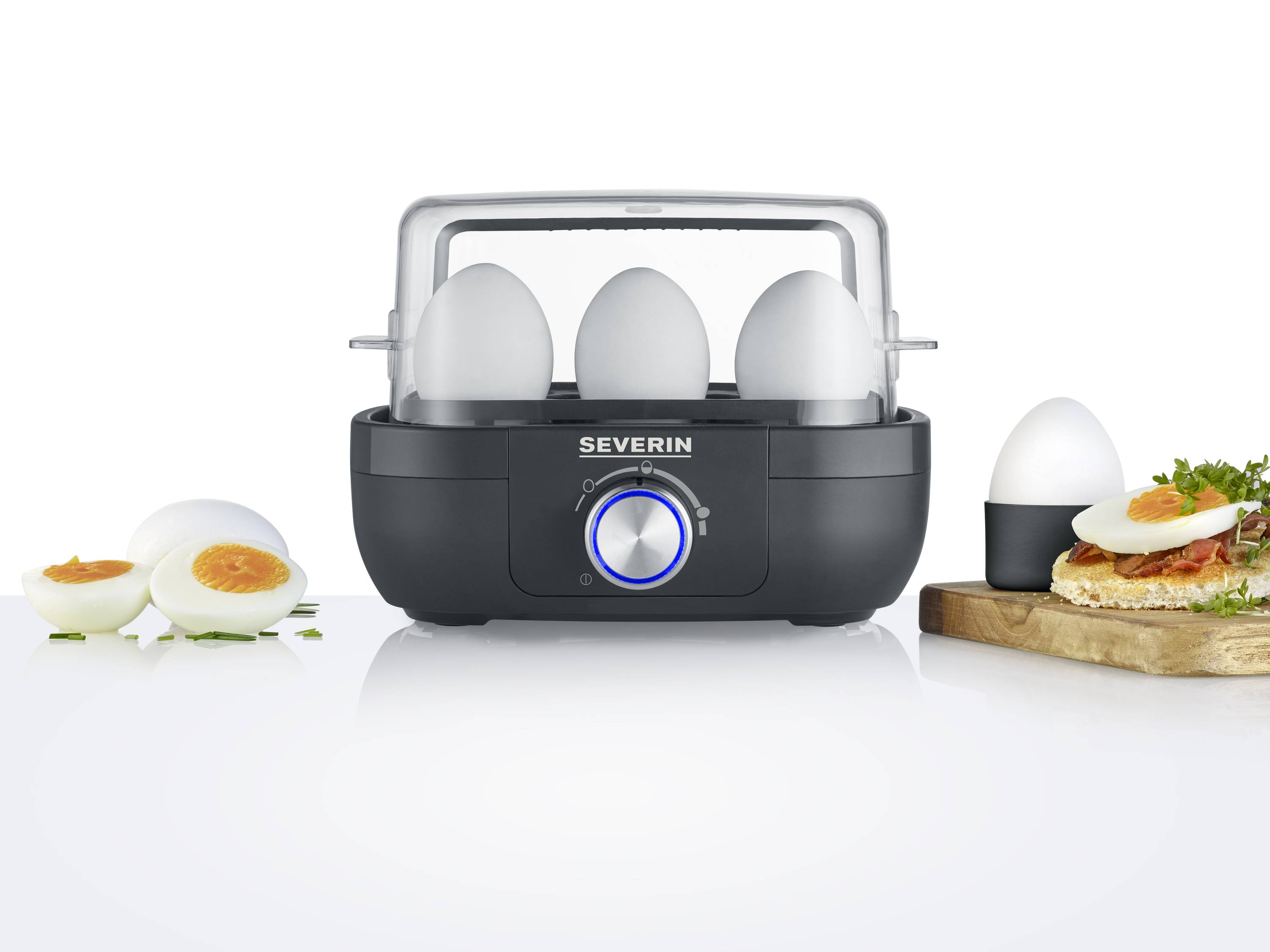 'Egg cooker with six eggs, three in the cooker and three on a table. Boiled eggs, sliced, on bread beside it.'