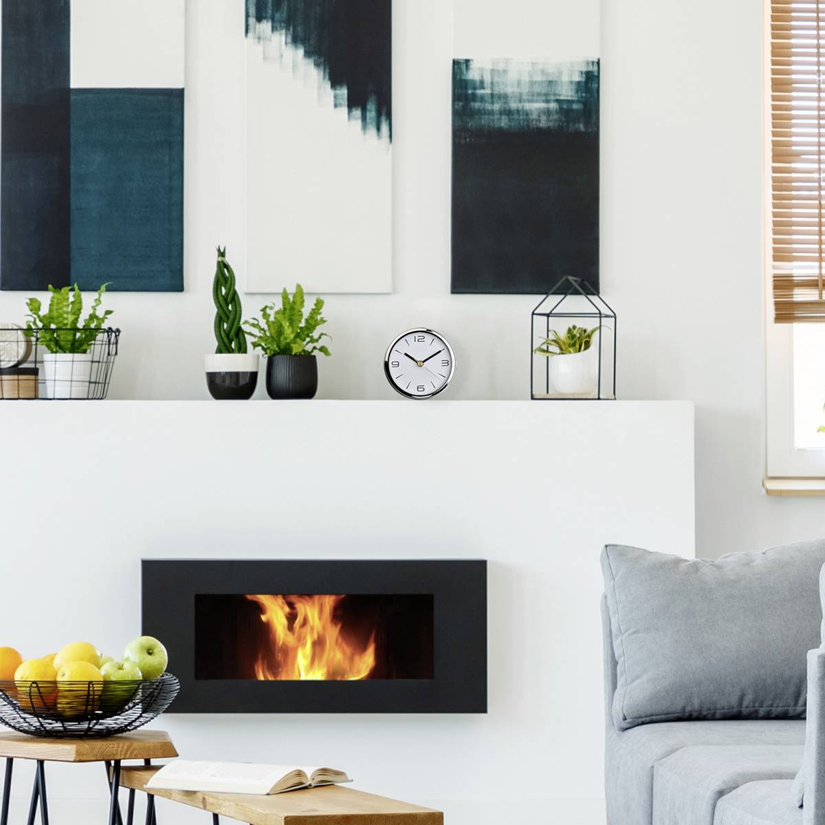 Living room with a modern fireplace, green plants, and wall paintings. A table with a fruit bowl stands in front of a grey sofa.
