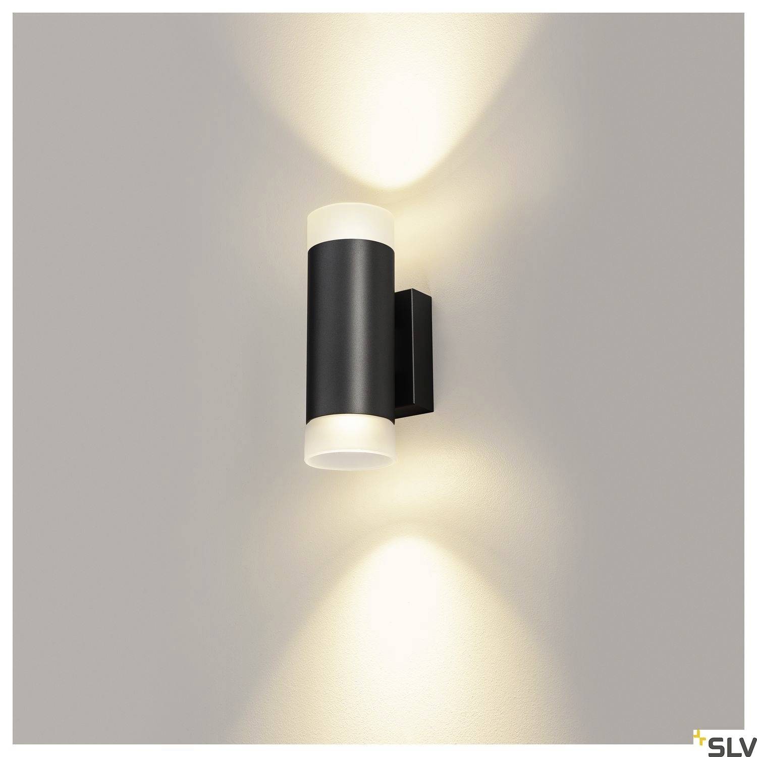 Black wall light with cylindrical design emits warm light upwards and downwards, mounted on a grey wall.