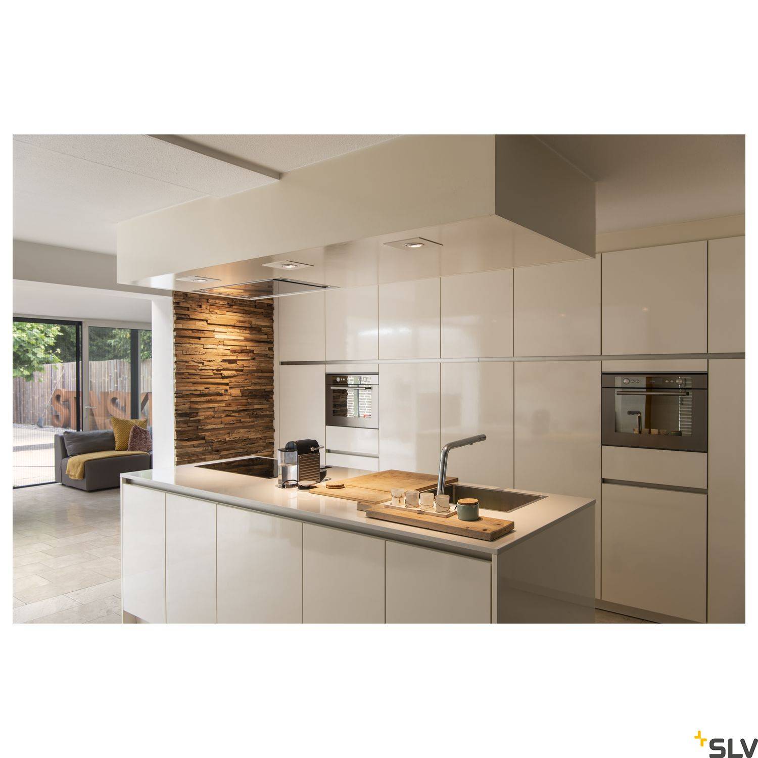 Modern kitchen with white high-gloss units, a central island hob and built-in oven. Window in the background shows garden view.