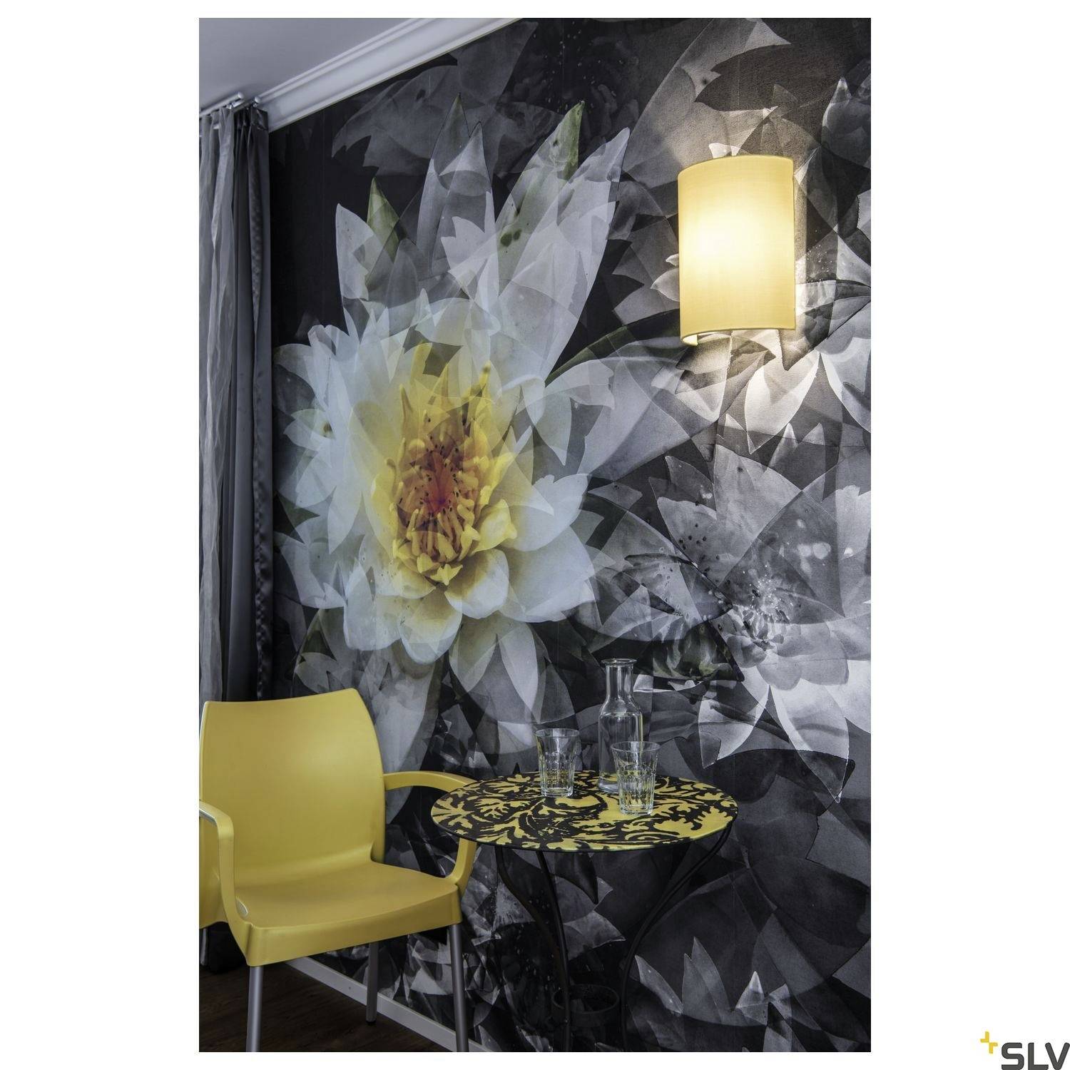 A yellow chair and a table with a floral design in front of a wall wallpaper with a large flower motif. A glowing lamp hangs on the wall.
