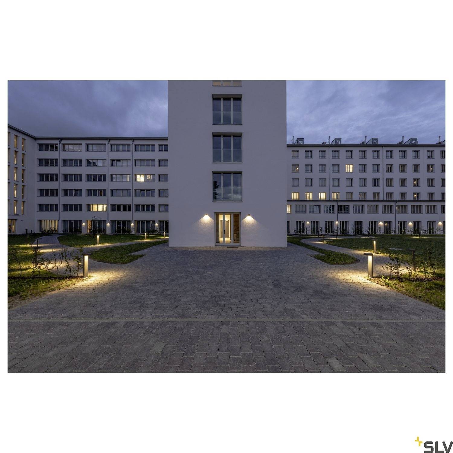 A lit entrance area of a multi-storey building at dusk, with symmetrically arranged pathway lights and well-maintained green spaces.