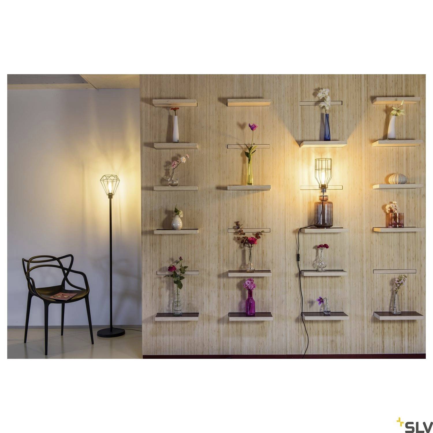 A wall with decorative flower vases on shelves, flanked by an illuminated standard lamp, creates a cosy atmosphere.
