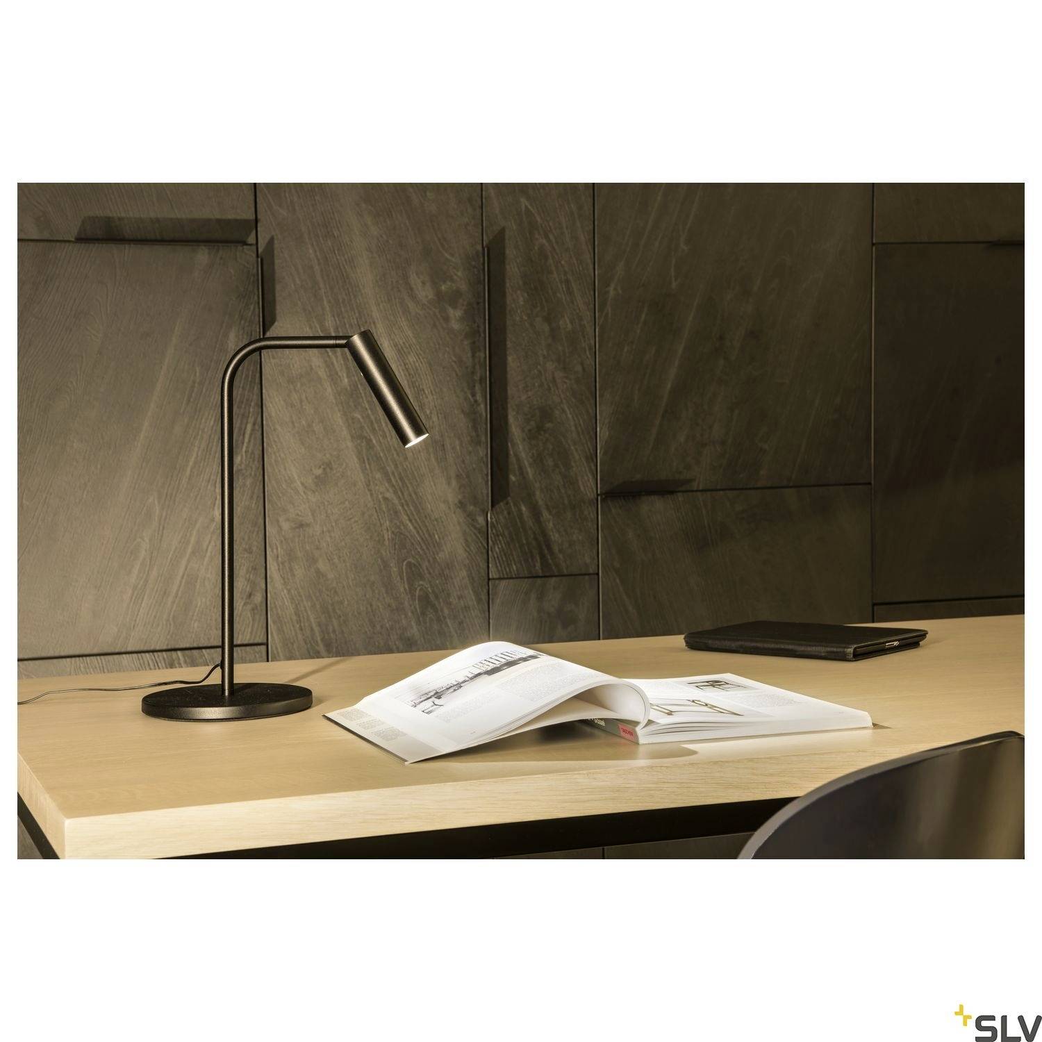 A black table lamp is sitting on a wooden table next to an open book and a closed laptop. A dark wall is visible in the background.