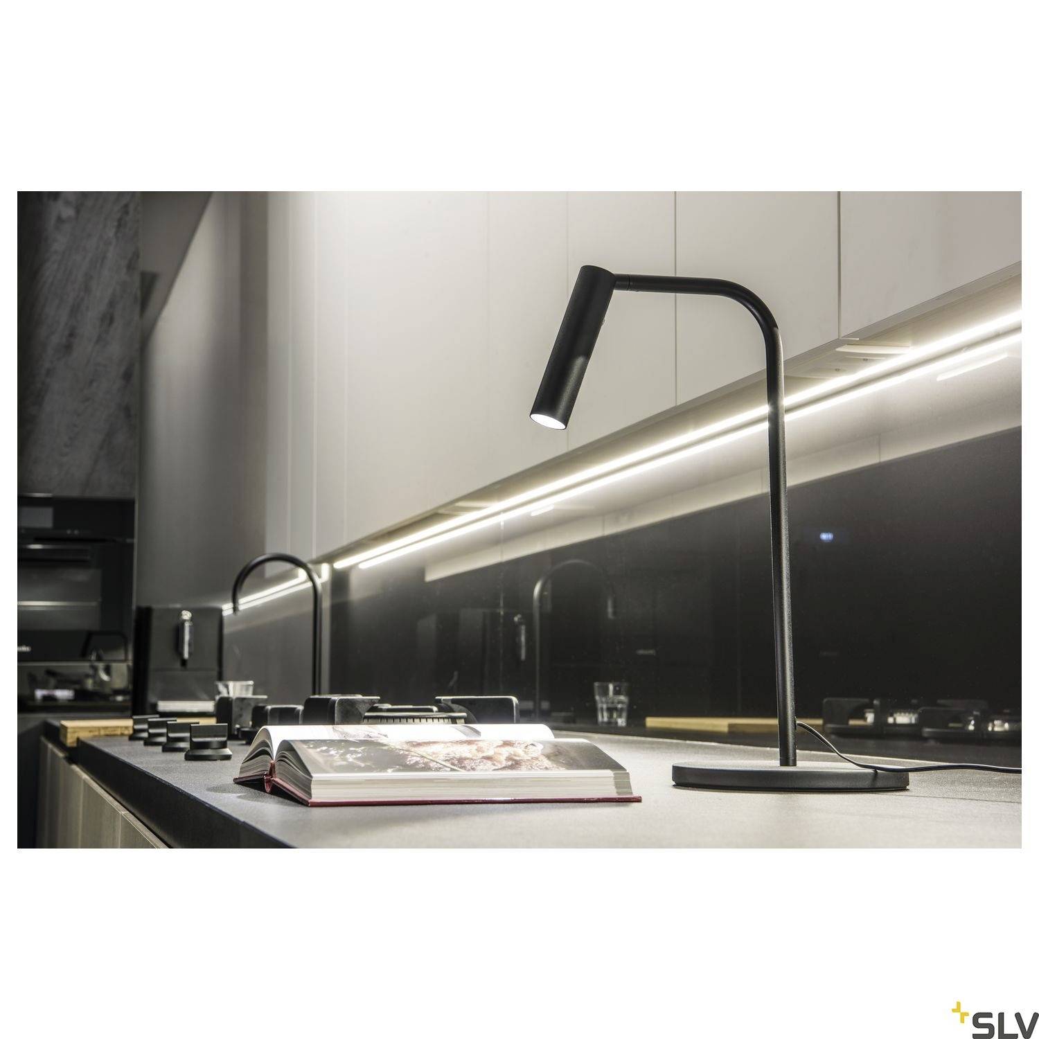 A modern kitchen with dark interior, illuminated by a sleek black table lamp. An open cookery book lies on the worktop.
