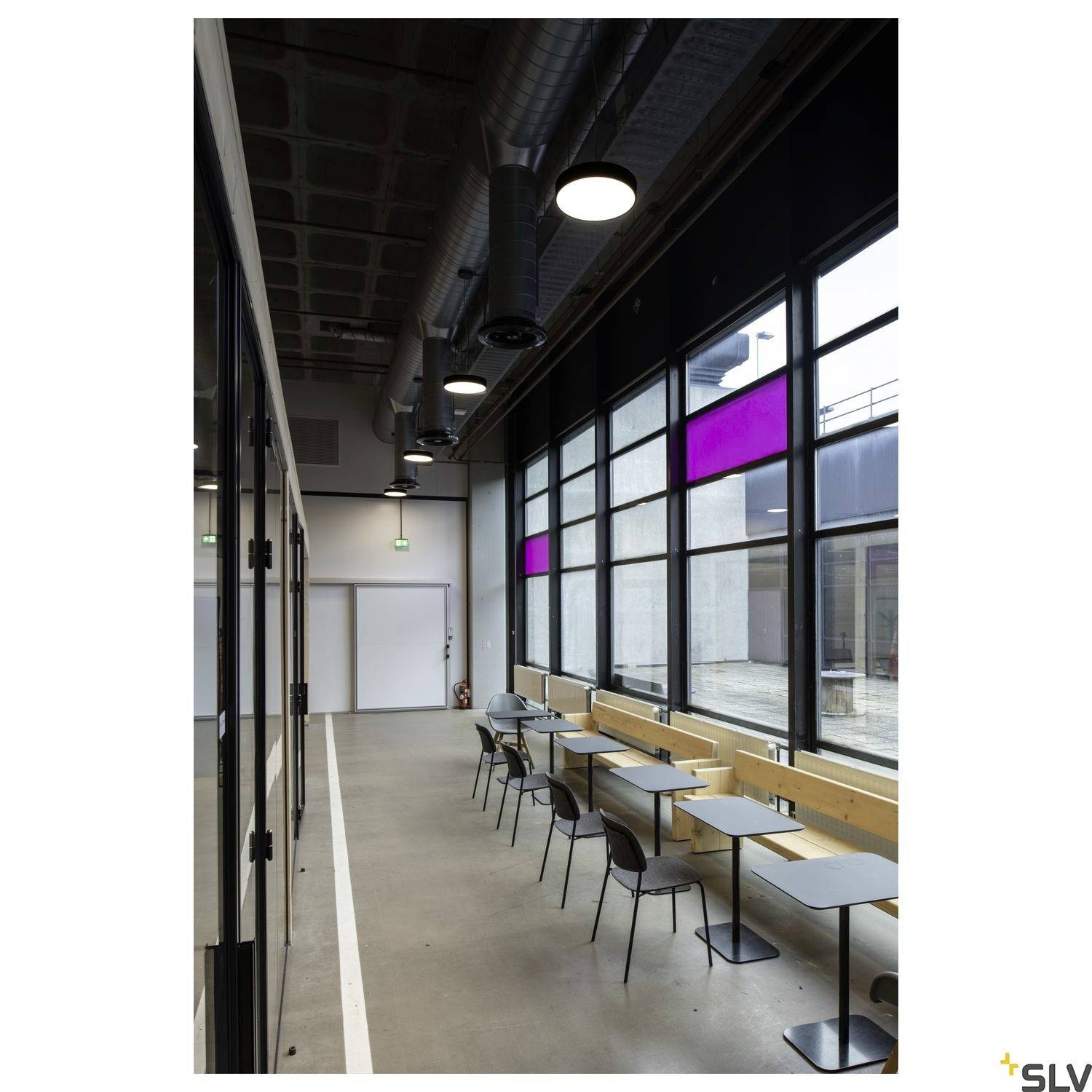 A modern interior with large windows and minimalist furnishings. Tables and chairs are positioned against the wall, and round pendant lights hang from the ceiling.
