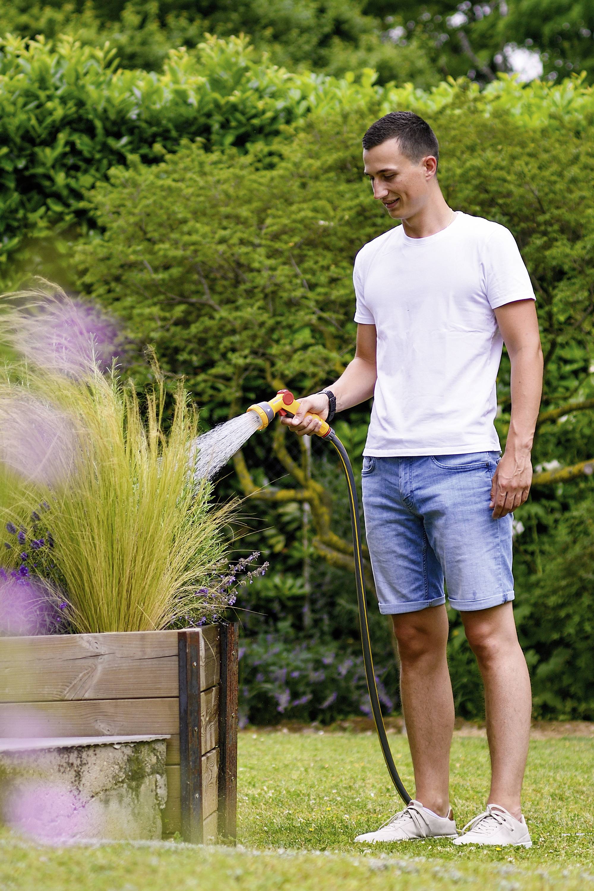 A man in a white T-shirt and denim shorts is watering plants in a wooden raised bed using a garden hose in a green garden.