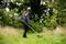 A person is mowing the lawn with a strimmer in a garden, surrounded by trees and tall grasses.