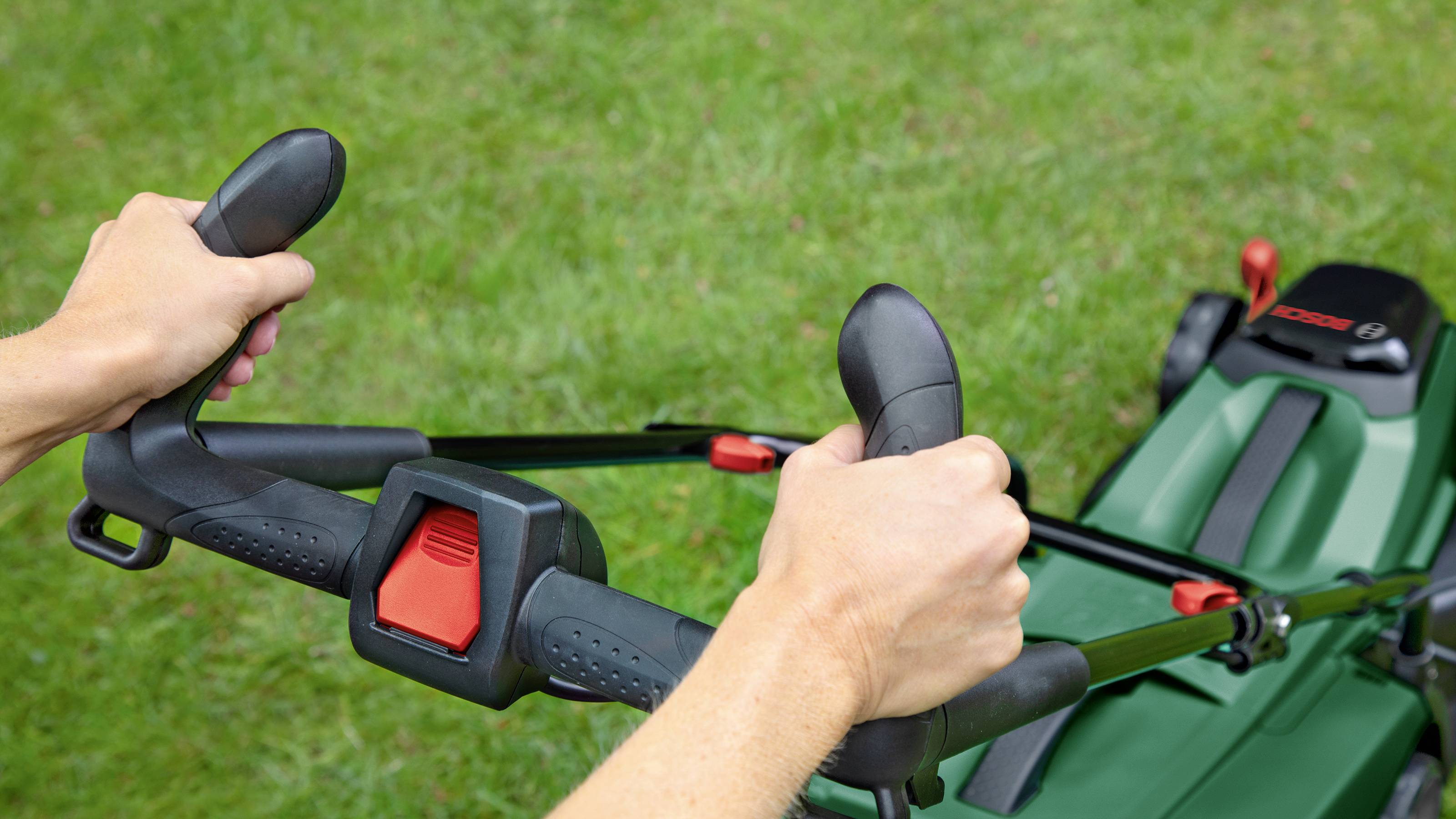 Hands grip the handles of a lawnmower on a green grassy area.