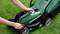 A person is adjusting the cutting height on a green lawnmower in a meadow. The lawnmower has black and red elements.