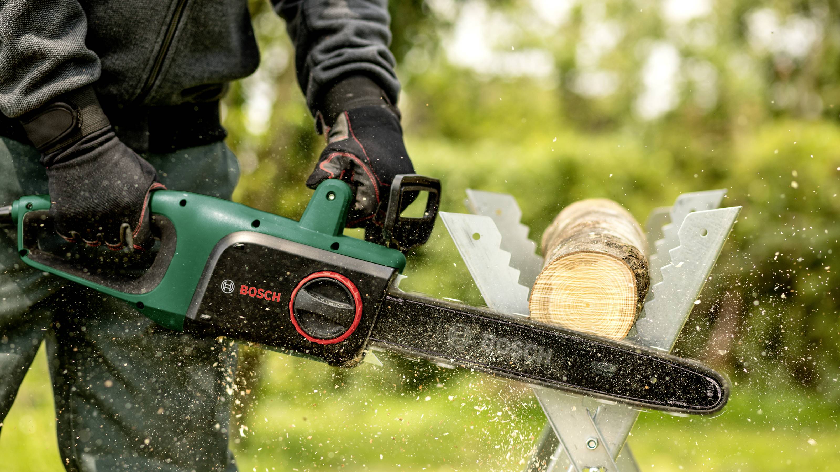 A person is sawing a log with an electric chainsaw on a sawing horse outdoors. Sawdust is flying around.