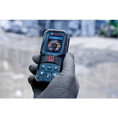 A hand is holding a Bosch laser distance measurer with display and controls. Background blurred, work scene.